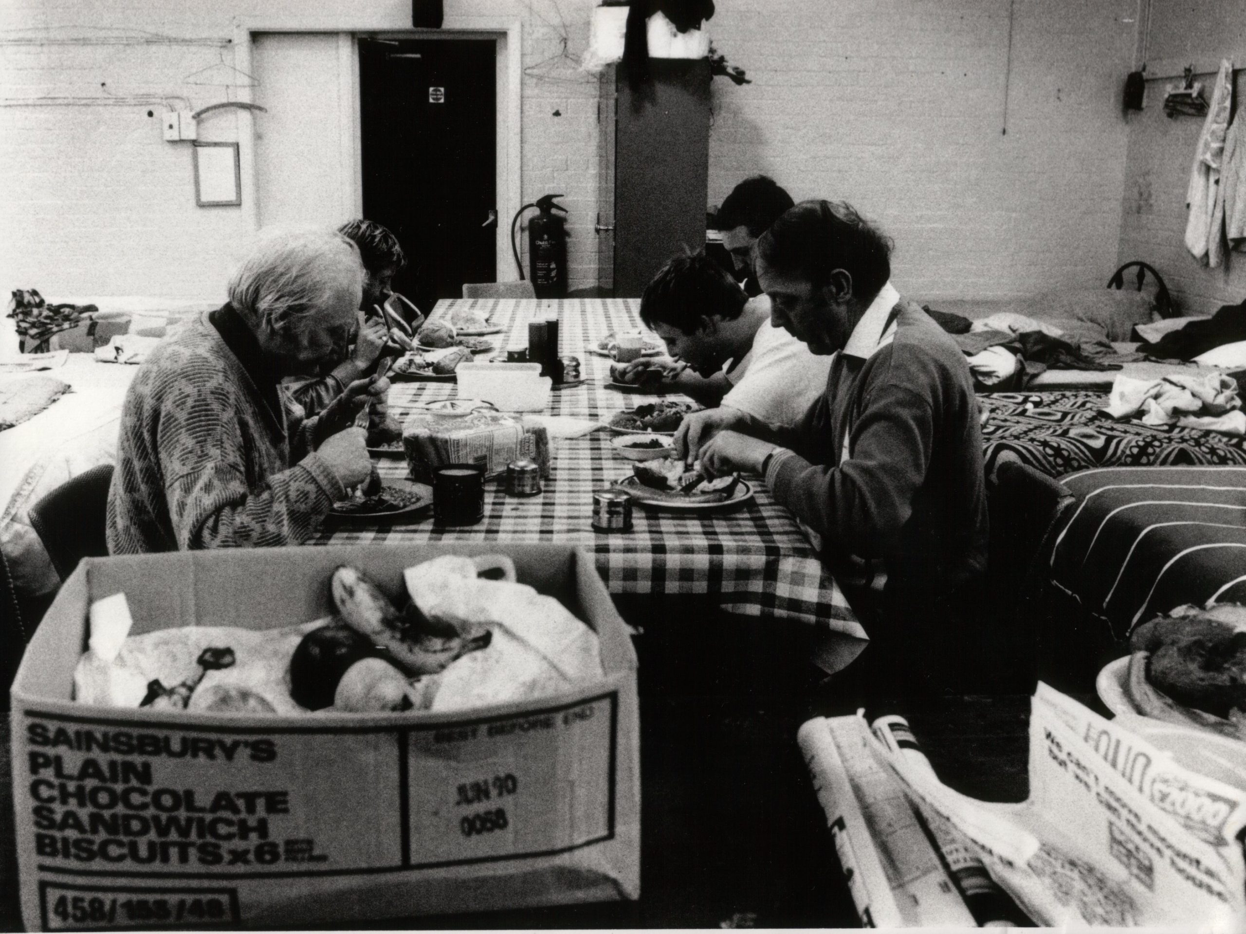 SSJ Our history image. Few homeless people sitting around a table eating, when The Society of St James was first established.
