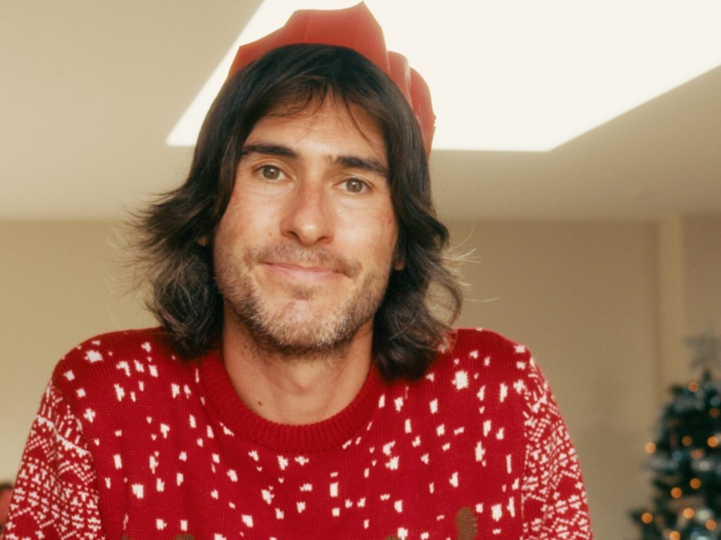 Joe, wearing a Christmas jumper and paper crown, smiling at home in front of a Christmas tree